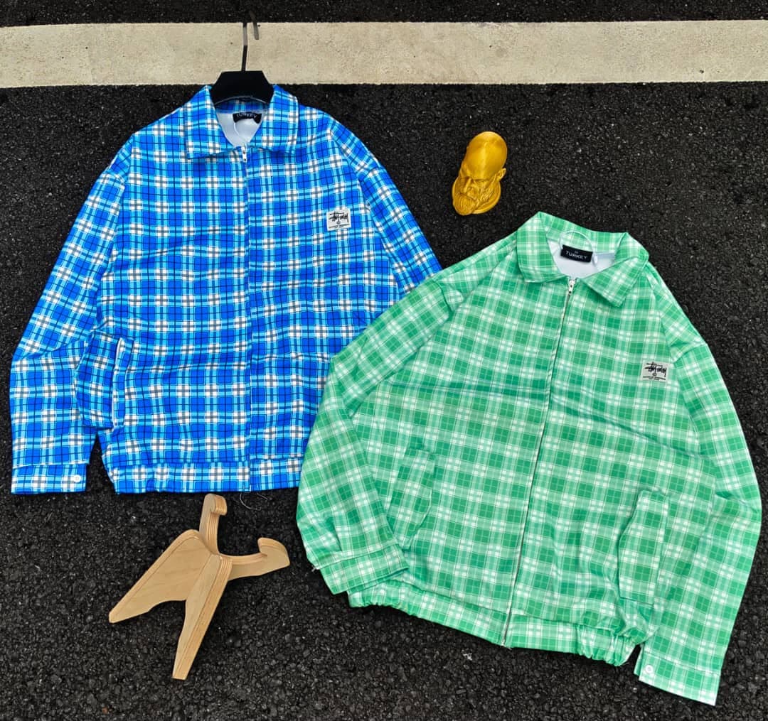 Blue and green Stussy plaid zip-up jackets laid out on asphalt with accessories.