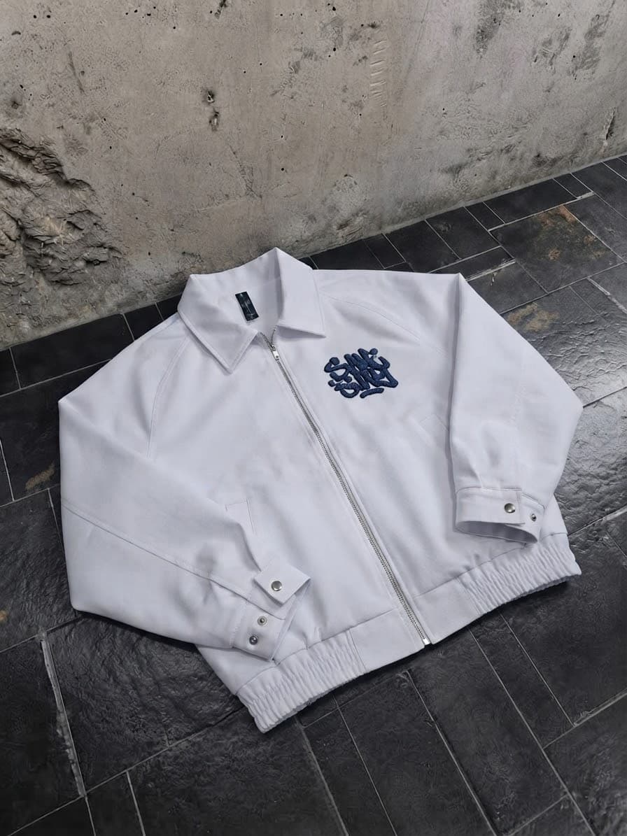 White zip-up bomber jacket with navy chest embroidery displayed on tiled floor.