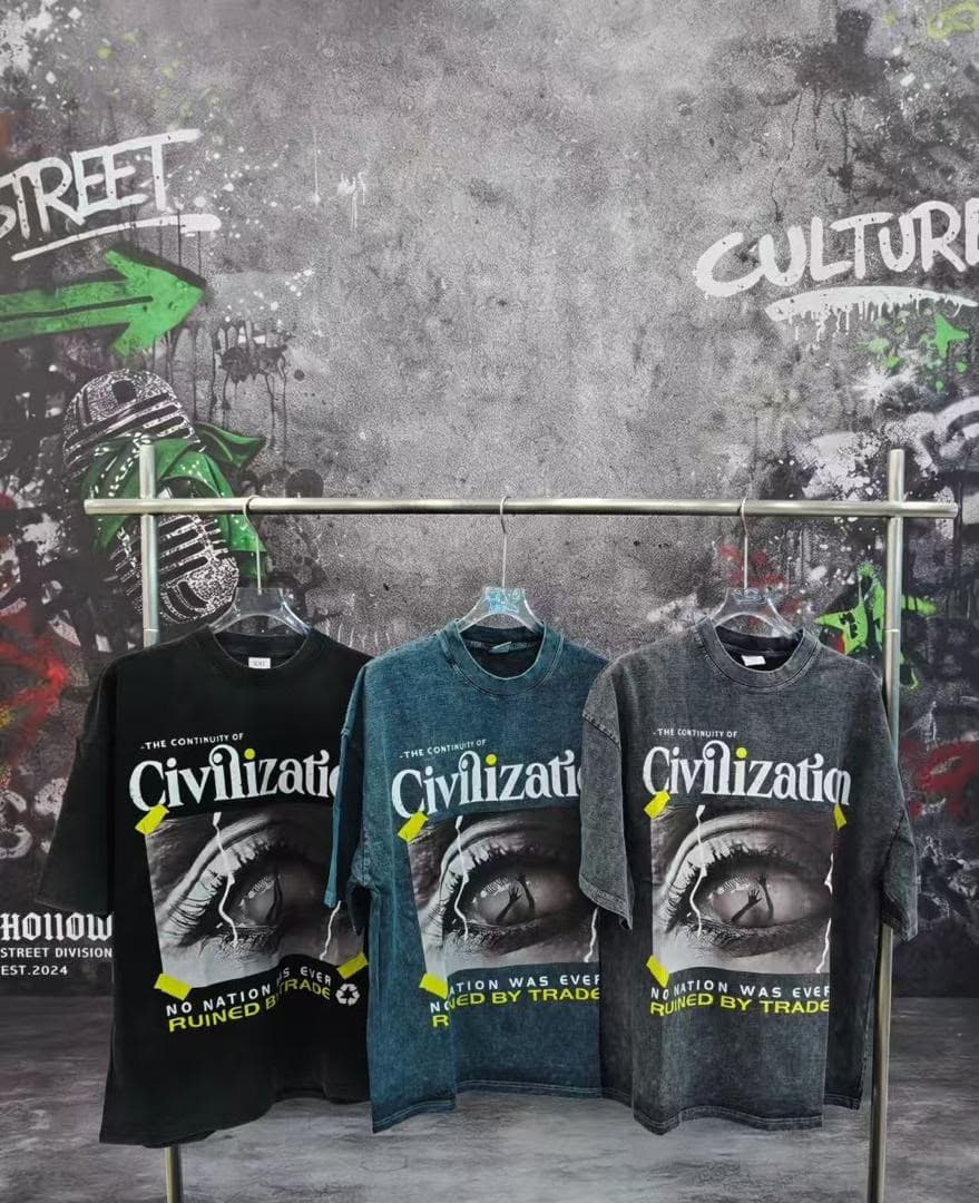 Black, teal, and gray stonewashed Civilization t-shirts with eye graphic hanging on rack against graffiti wall.