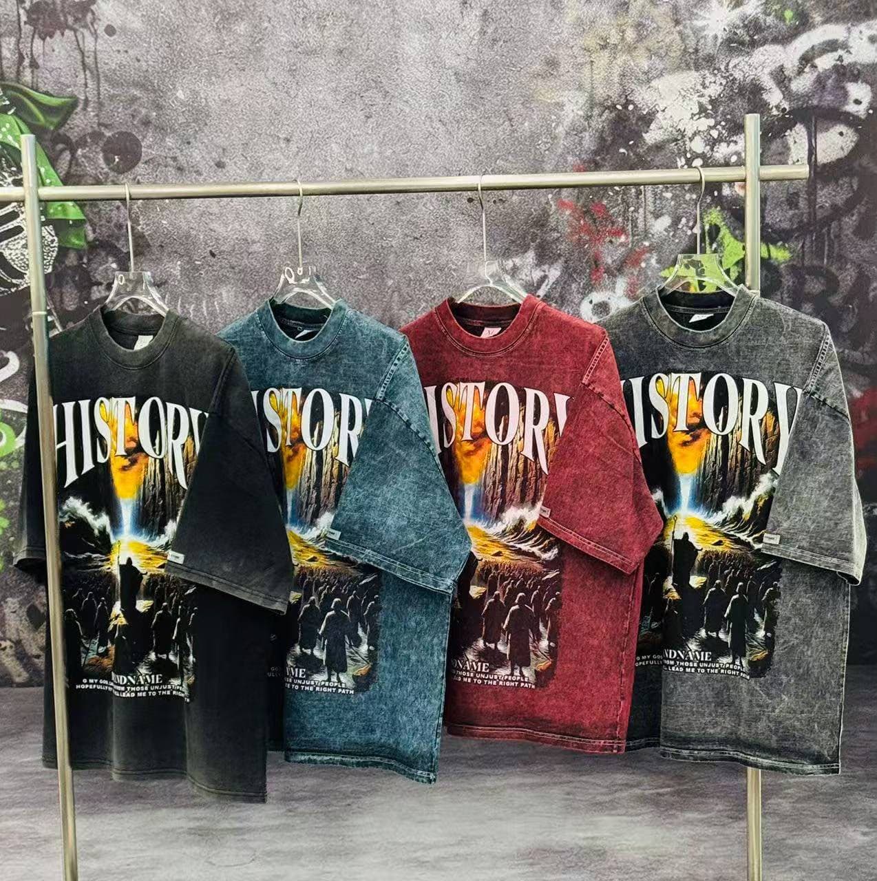 Four stonewashed History graphic t-shirts in black, blue, red, and gray hanging on metal rack with graffiti backdrop.