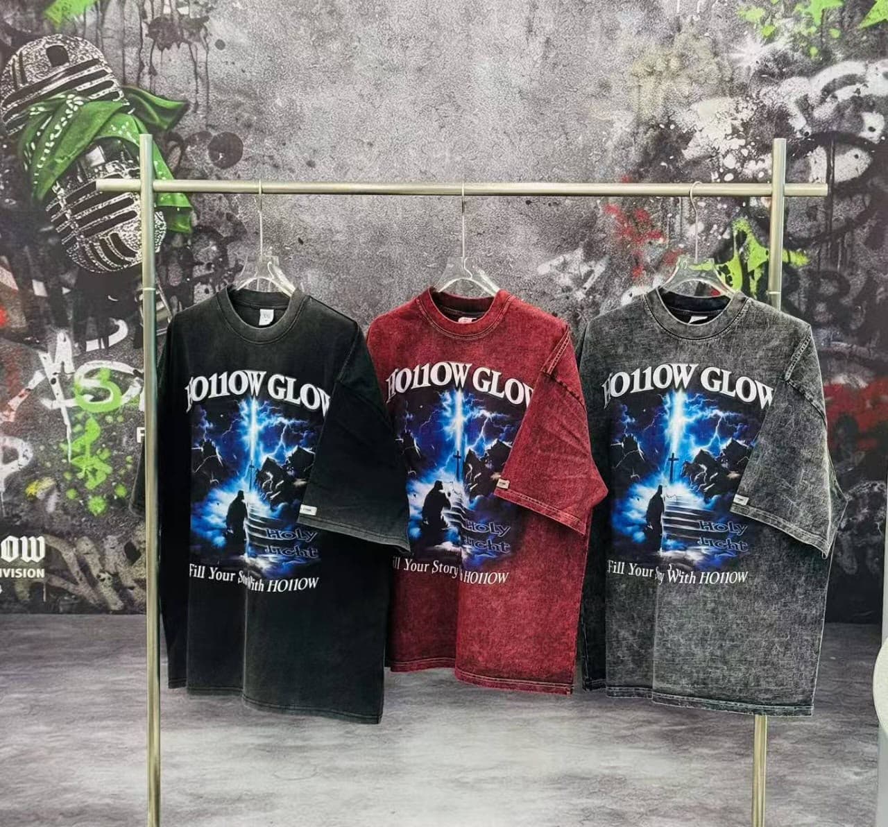 Black, red, and gray Hollow Glow graphic T-shirts hanging side by side on a clothing rack against graffiti wall.