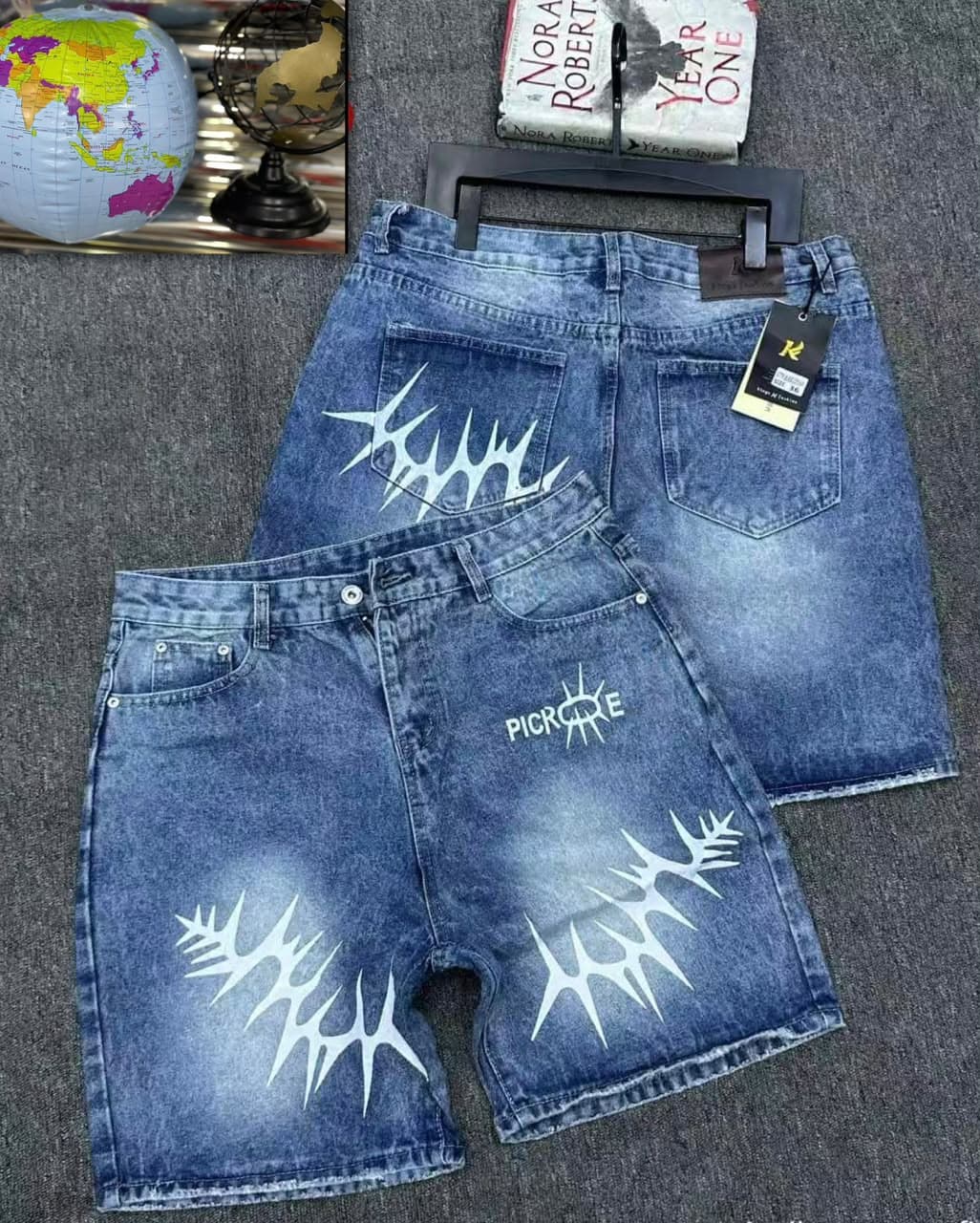 Blue denim shorts with white spike prints, PICRONE graphic, and back views on grey surface with book and globe props.