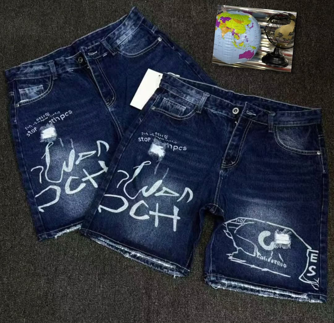Blue denim shorts with white graffiti prints, text, and distressed effects laid flat on dark background with globe inset.