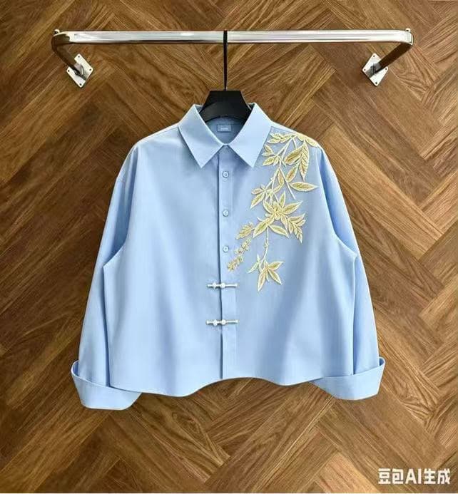 Light blue cropped button-up shirt with gold leaf embroidery on wooden background.