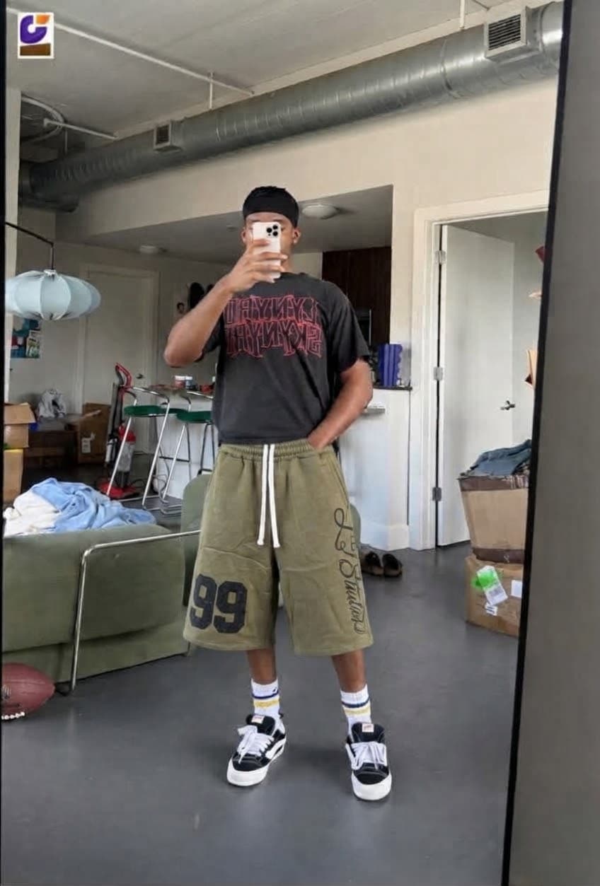 Man sitting in studio wearing black 99 print jorts with black hoodie and slides.