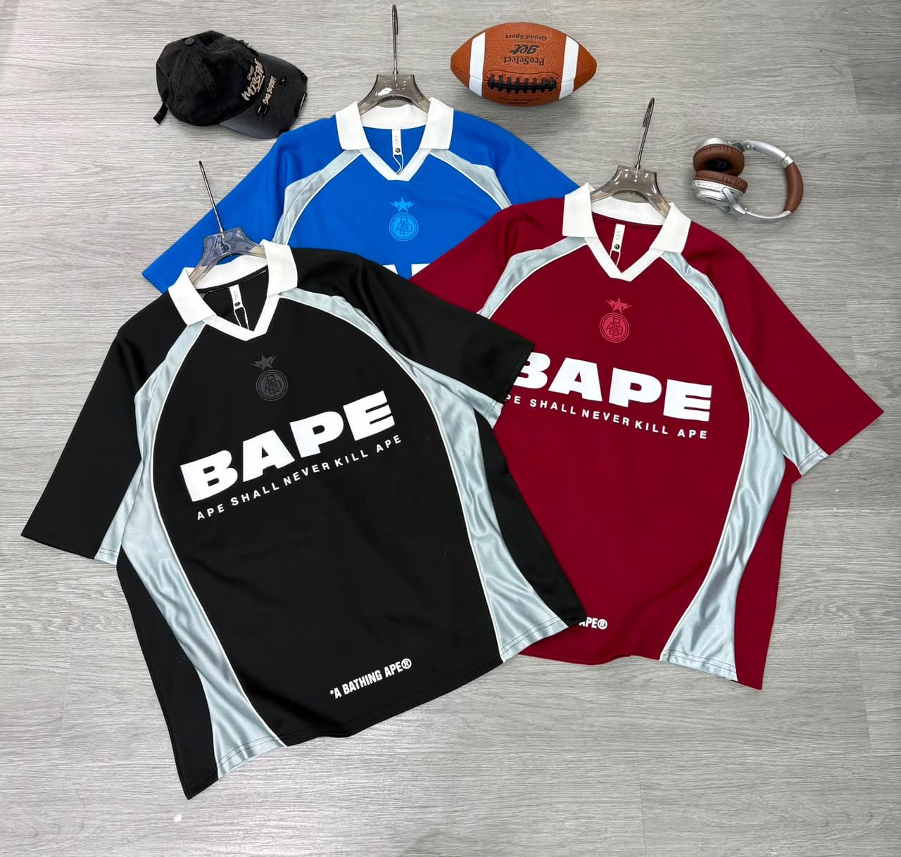 Flat lay of black, blue and red BAPE jersey t-shirts with cap, football and headphones.