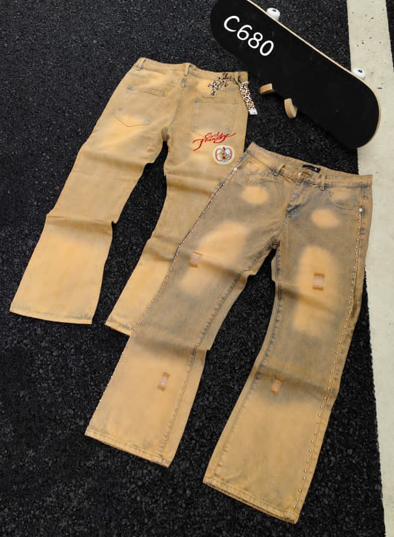 Two views of tan distressed baggy jeans with embroidery, studs and skateboard prop on dark pavement.