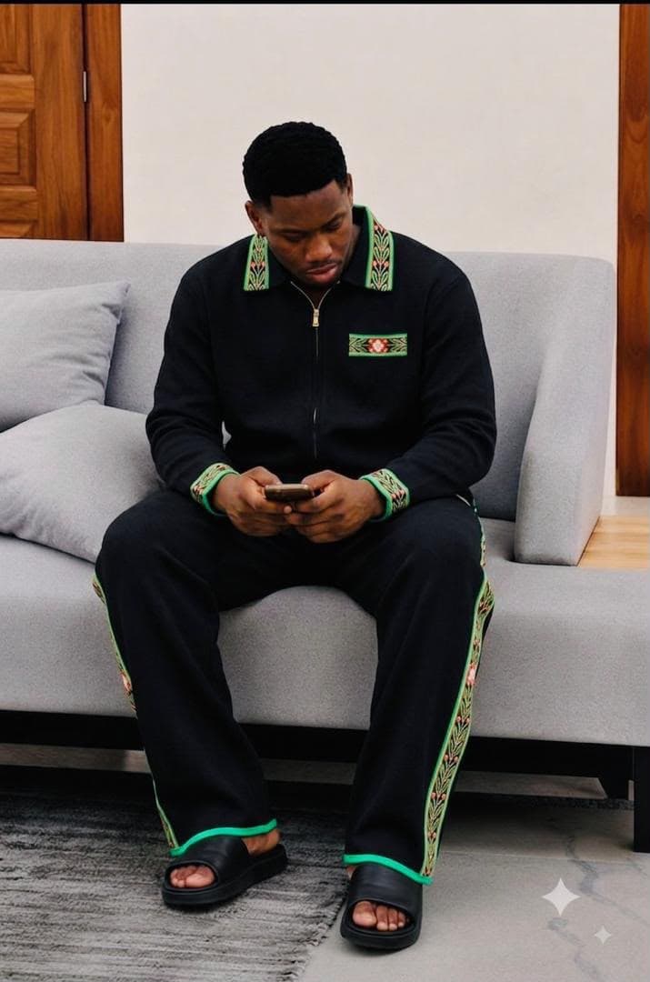 Man seated on couch wearing black embroidered zip-up tracksuit with black slides.