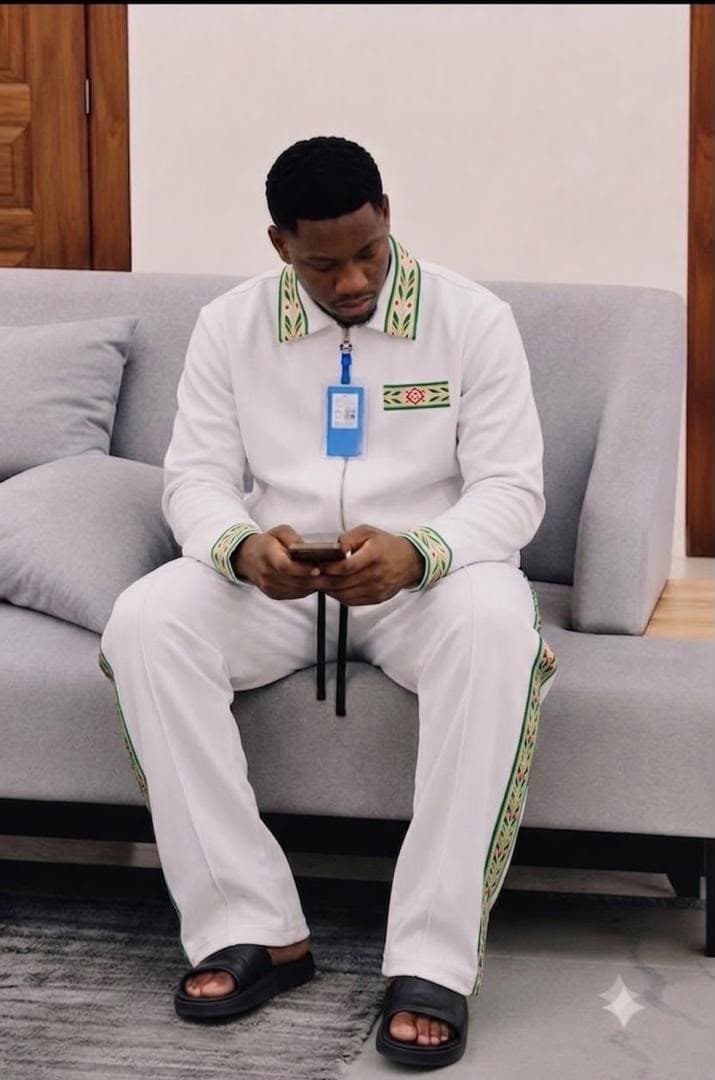 Man seated on couch wearing white embroidered zip-up tracksuit with black slides.