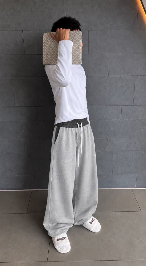Model wearing gray wide-leg joggers with white long-sleeve top and slippers, holding patterned clutch over face.