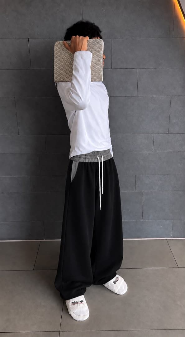 Model wearing black wide-leg joggers with white long-sleeve top and slippers, holding patterned clutch over face.