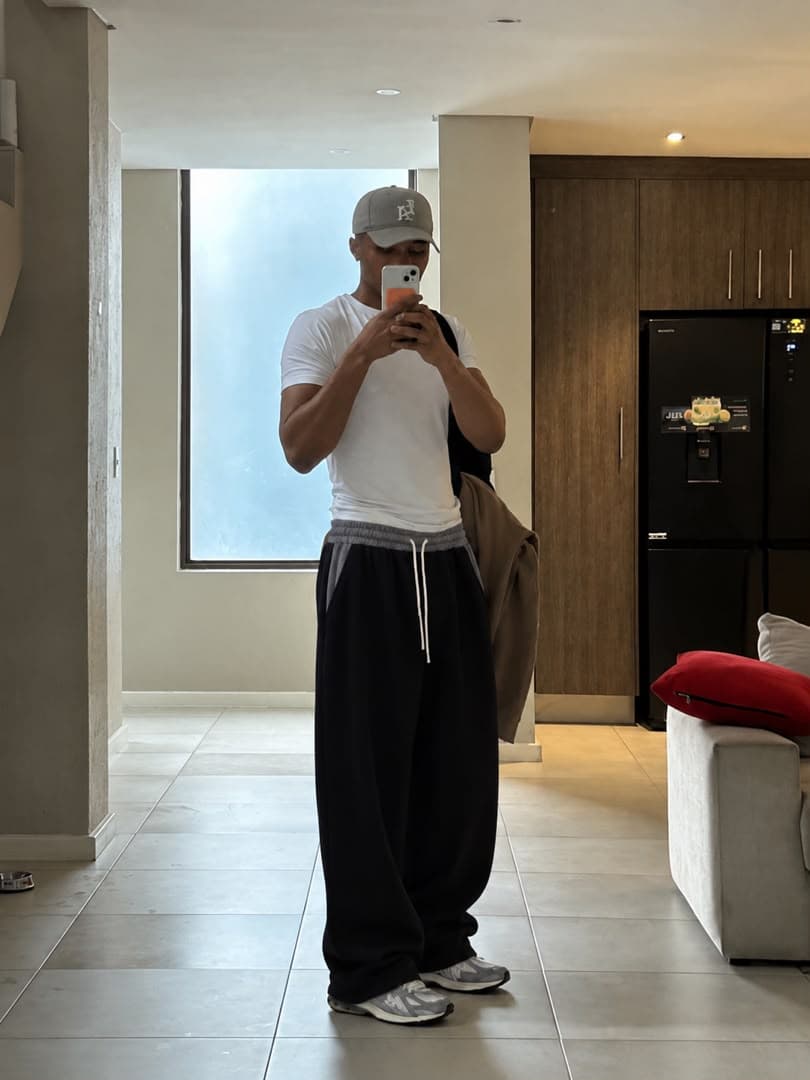 Man in white t-shirt and black wide-leg joggers taking mirror selfie in living room.
