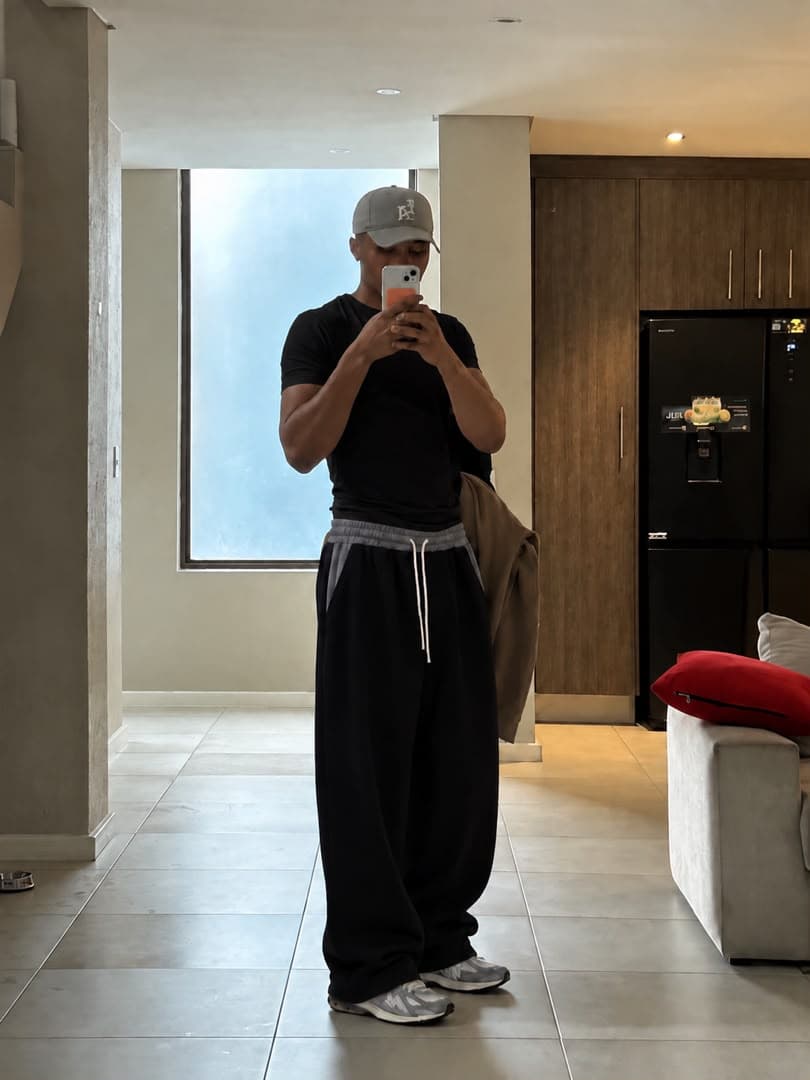 Man wearing black wide-leg joggers, black t-shirt and cap taking selfie in modern living room.