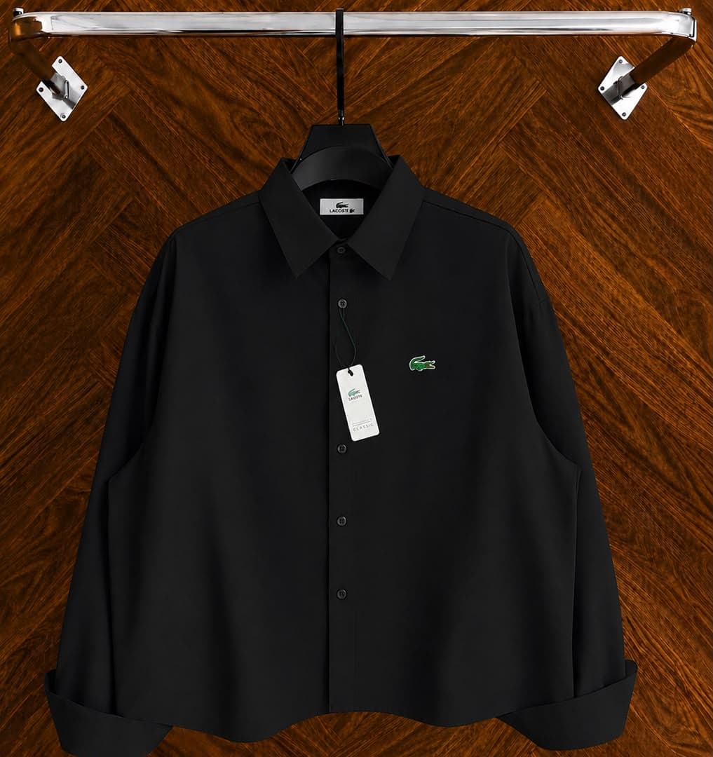Black Lacoste long sleeve button-down shirt with signature green croc logo hanging against wood floor.