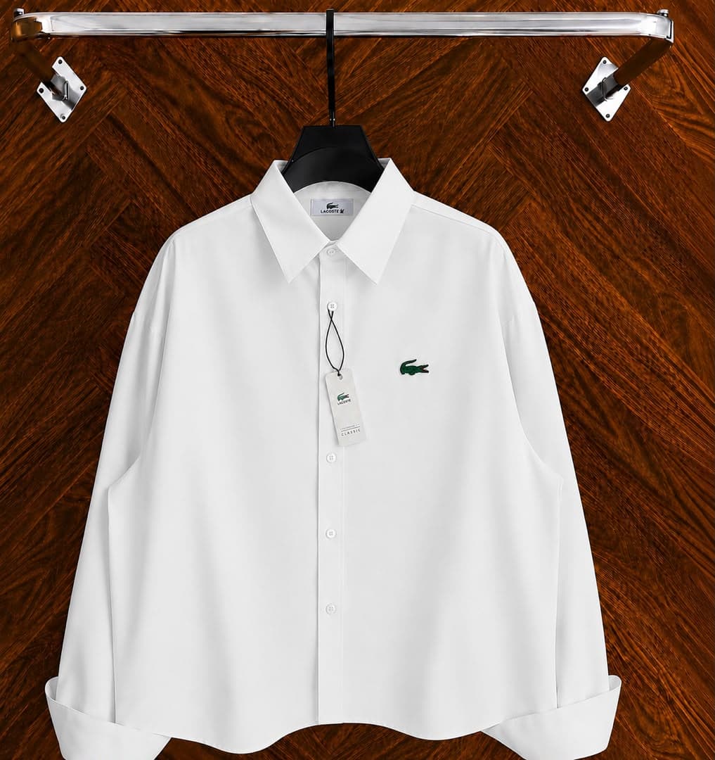 White Lacoste long sleeve button-down shirt with green croc logo on hanger against wooden background.