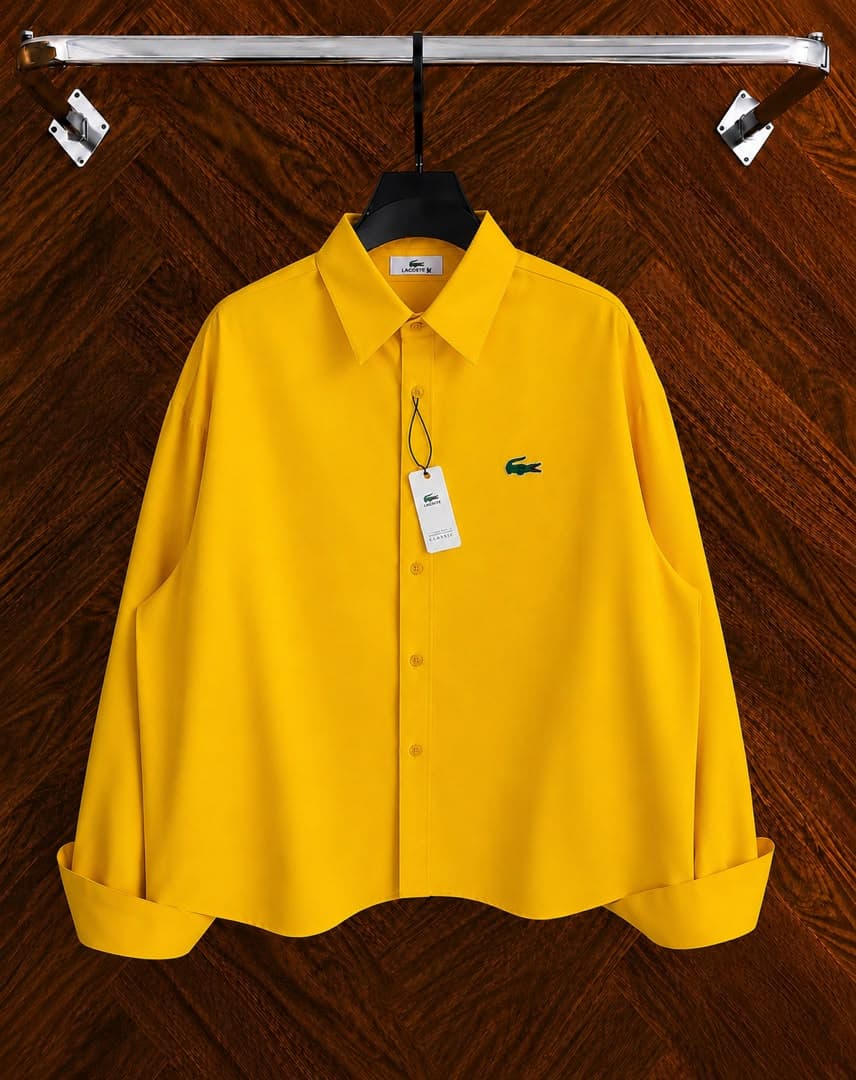 Yellow Lacoste long sleeve button-down shirt with croc logo hanging on a black hanger against wooden floor.
