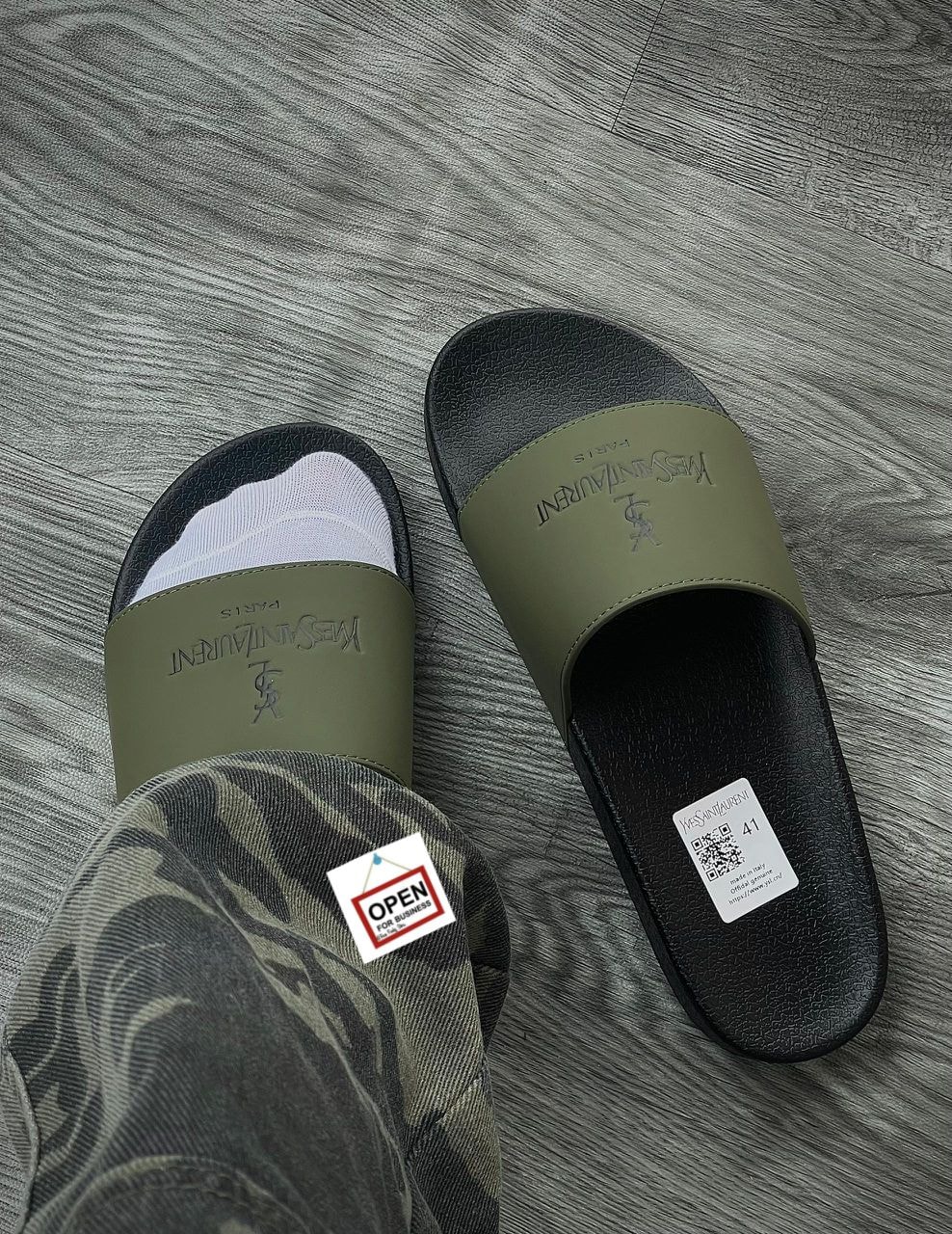 Person wearing olive YSL slide with camouflage pants on grey wood floor.
