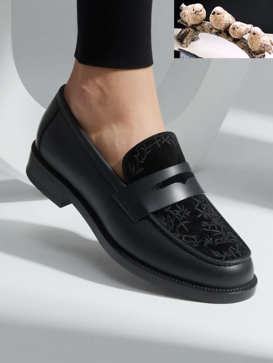 Black loafer with embroidered suede panel and metal bar worn on foot in studio setting.