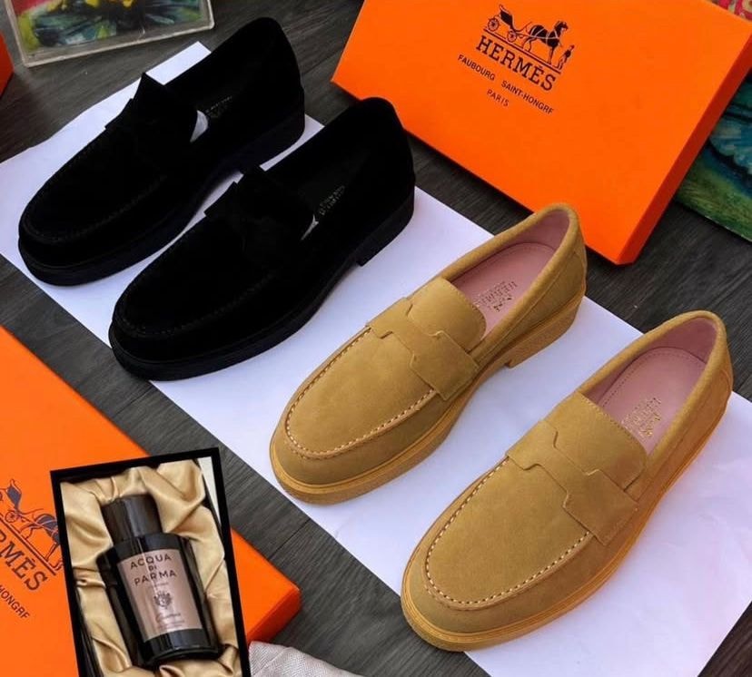 Black and tan suede loafers displayed alongside orange Hermes boxes, perfume and accessories.