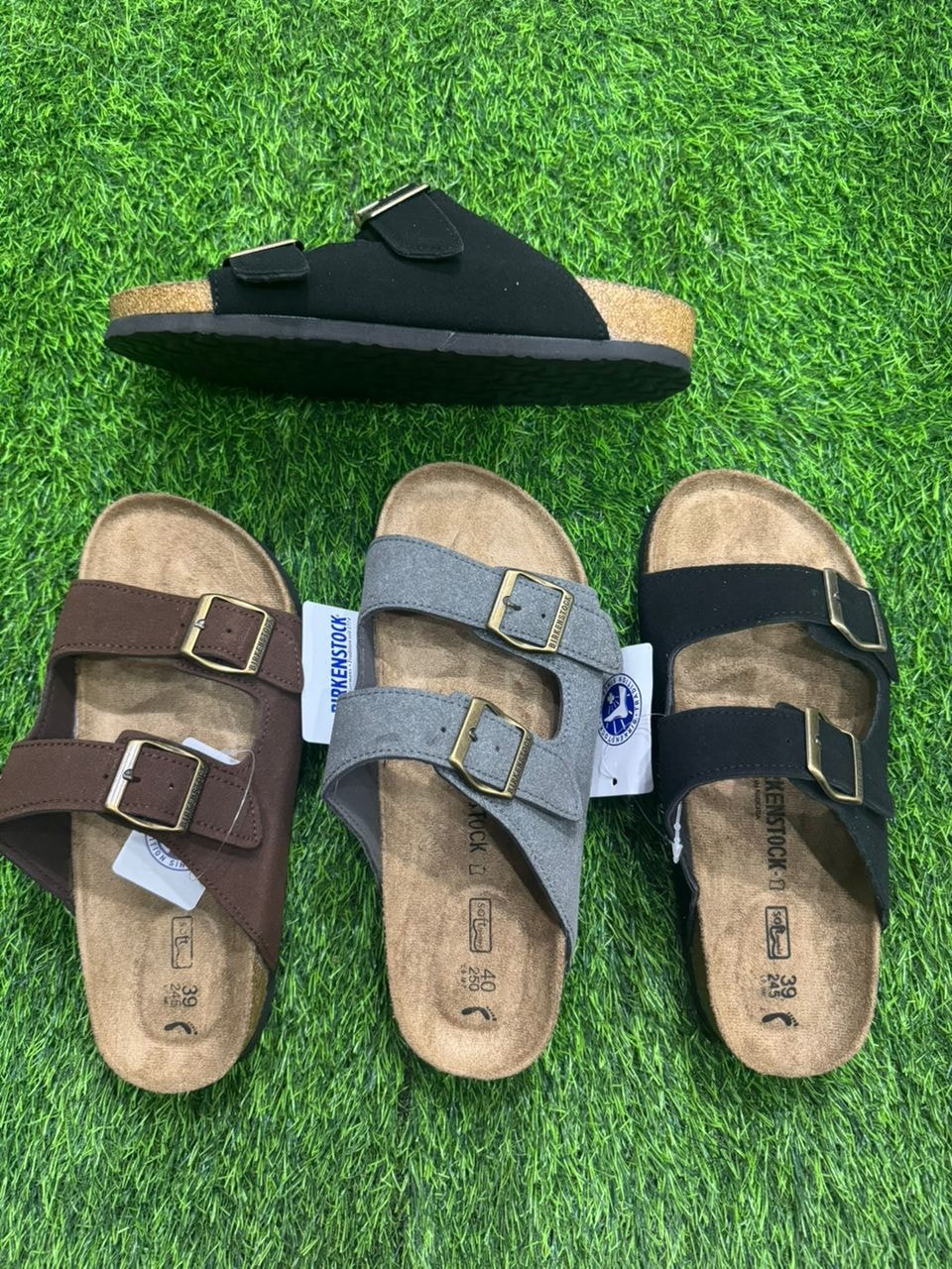 Birkenstock two-strap slides in brown, gray, and black shown from top and side angles on green grass.