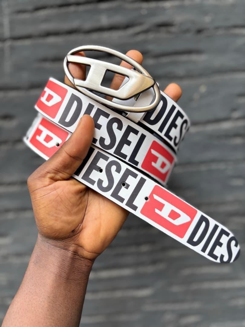 Hand holding a white Diesel logo belt with silver oval buckle against a brick wall.