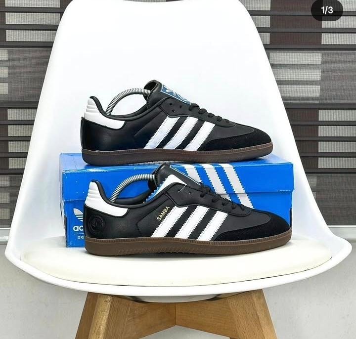 Black Adidas Samba sneaker with white stripes, leather and suede panels, displayed on chair with blue Adidas box.