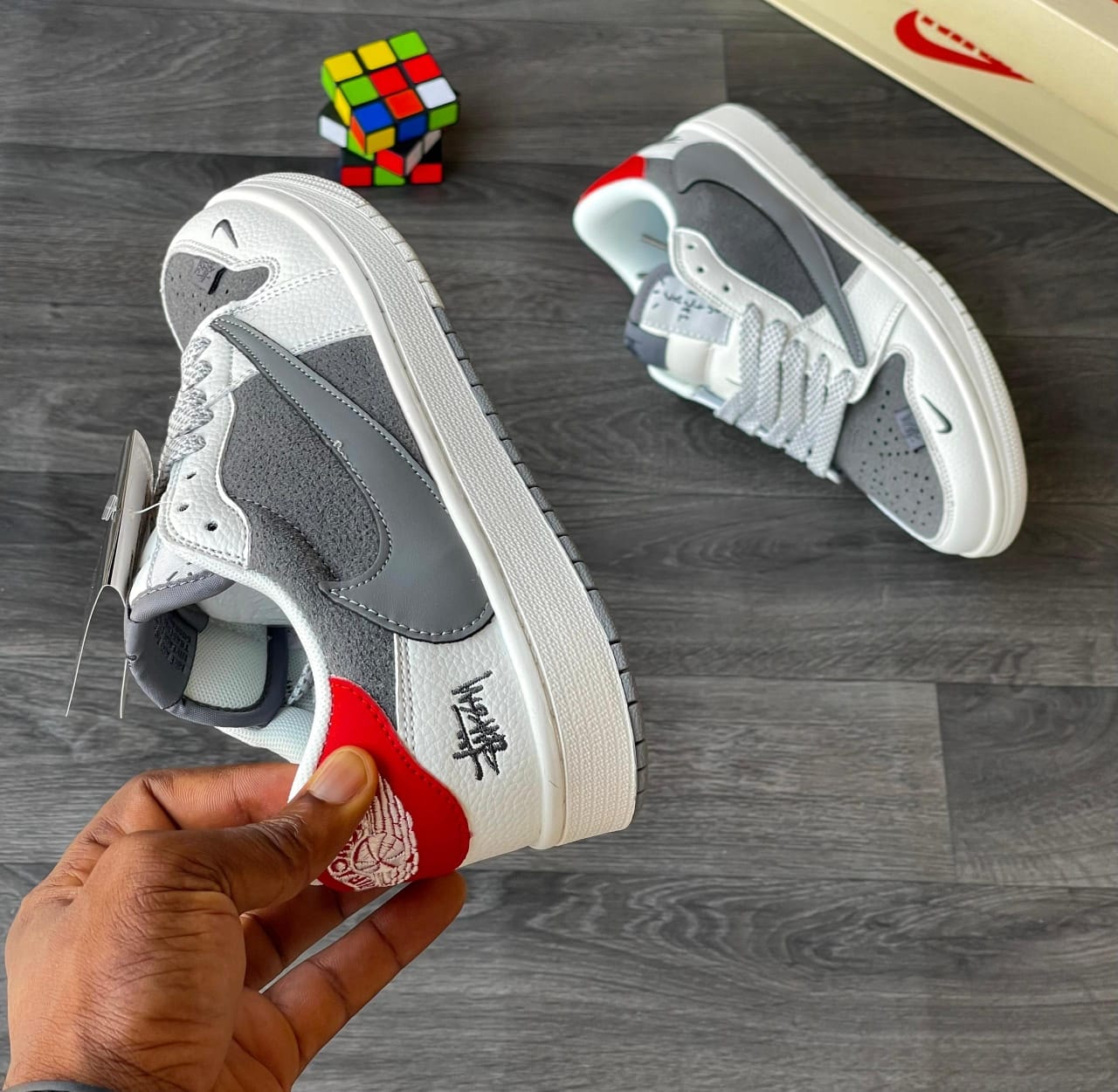 Hand holding grey Nike x Stüssy Air Force 1 sneaker with another pair and Rubik's cube in background.