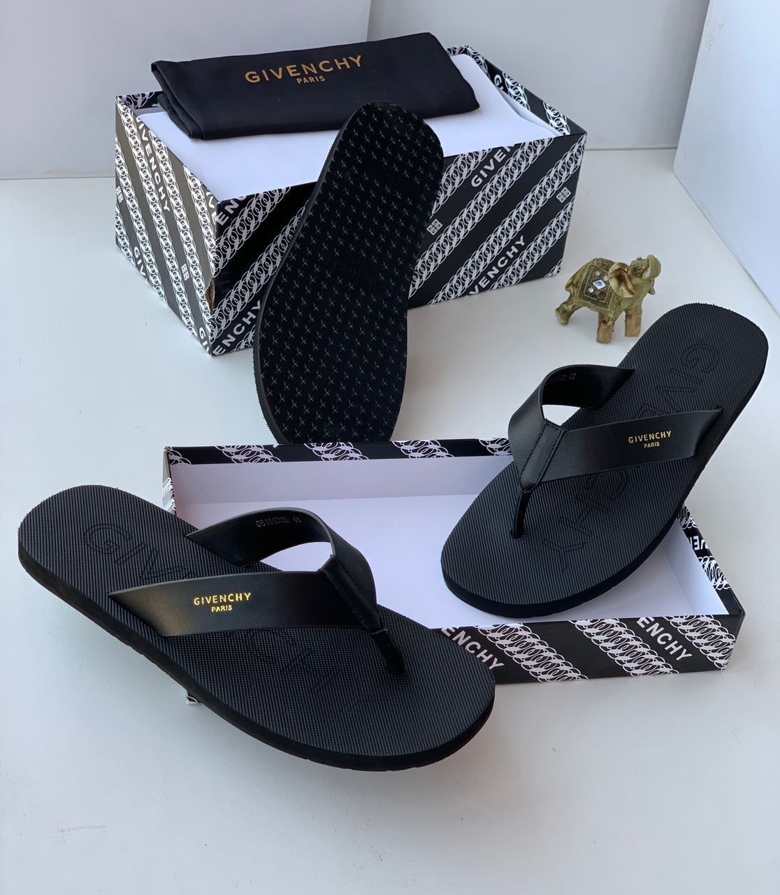 Black Givenchy flip flops shown with patterned box, dustbag and gold elephant figurine.