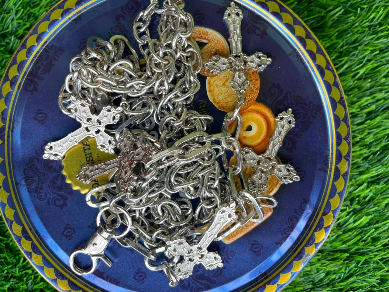 Pile of silver jean chains with ornate cross pendants on blue plate over grass.