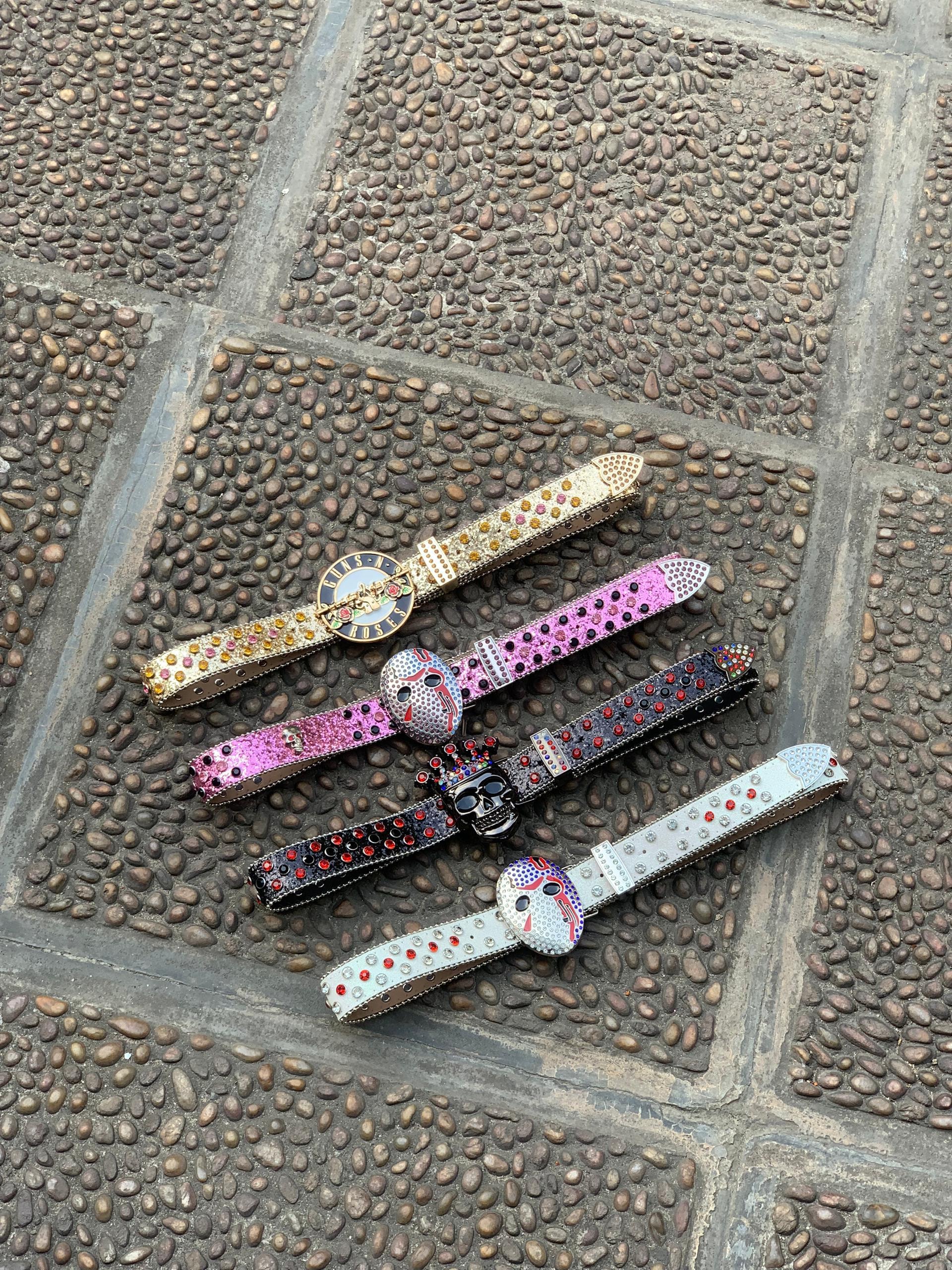 Four jeweled belts with skull and Guns N Roses buckles in gold, pink, black and white on cobblestone.