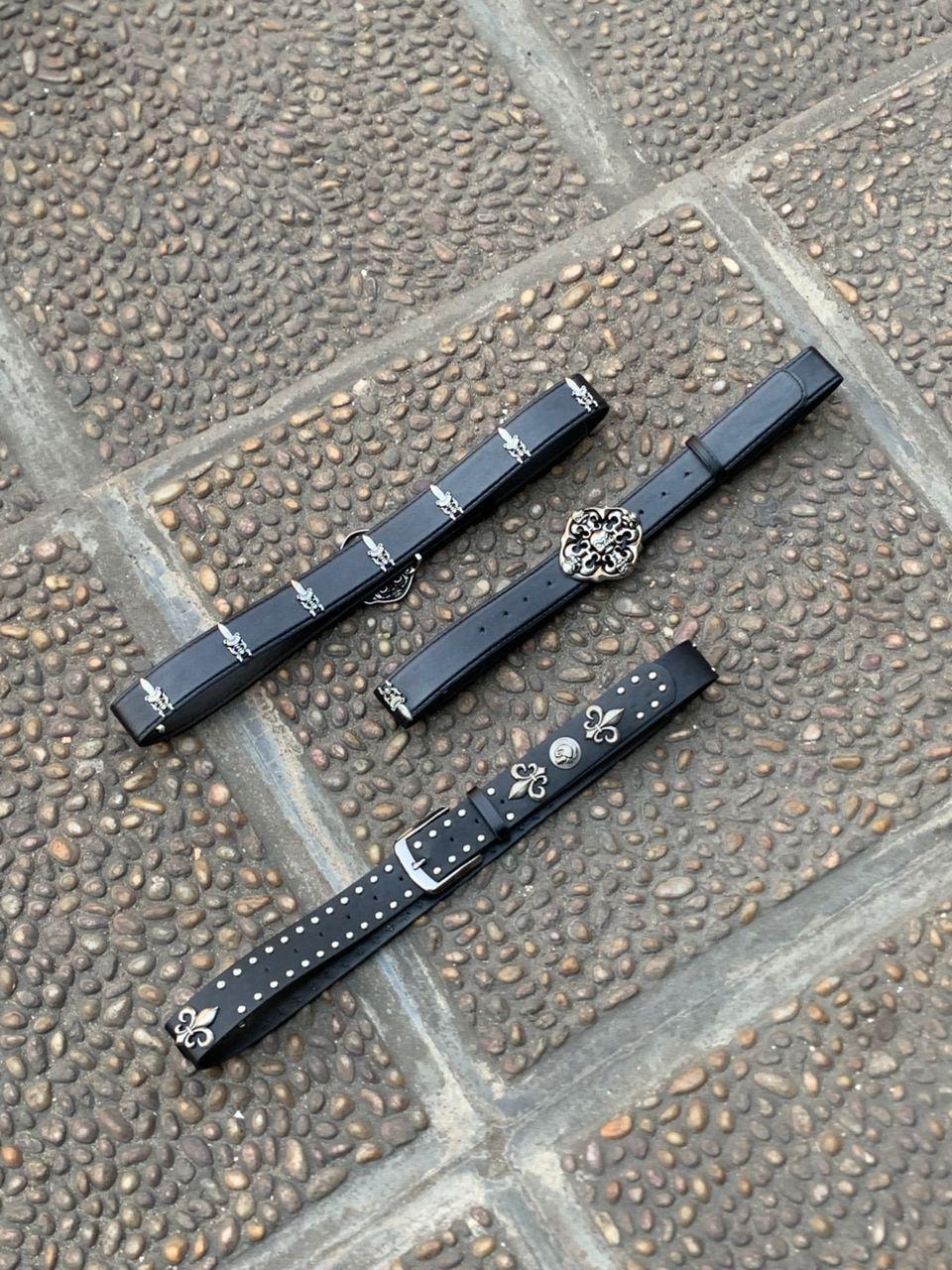 Three black leather belts with fleur-de-lis designs, studs, and various ornate buckles on a paved surface.