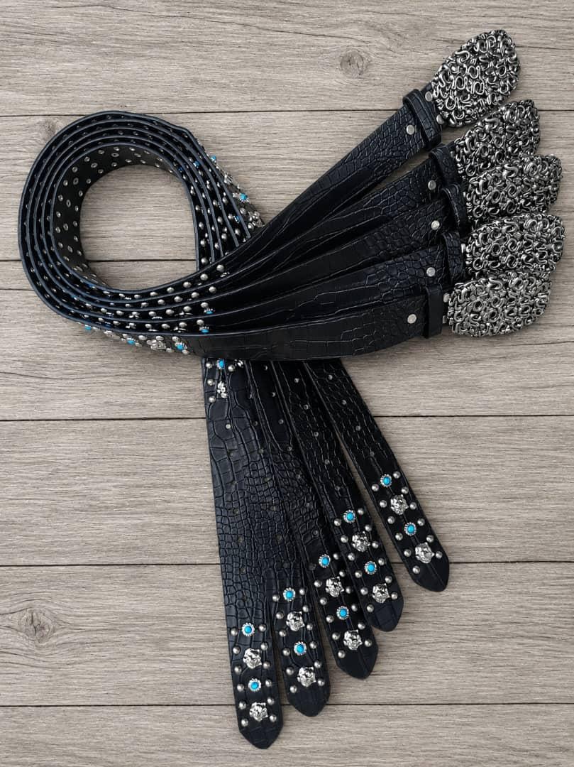 Collection of black designer belts featuring turquoise accents, studs and ornate silver buckles on a wooden surface.