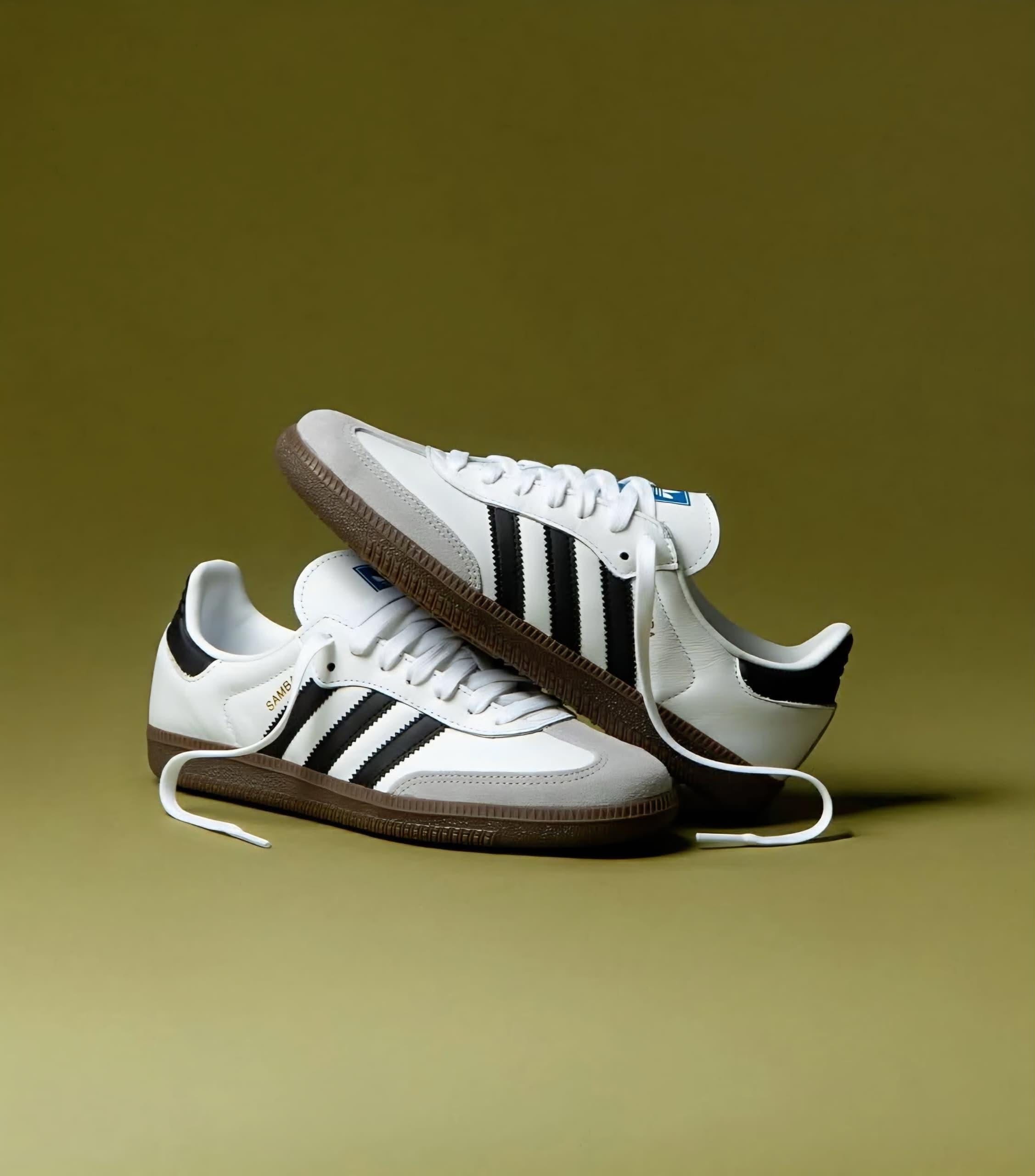 White Adidas Samba sneakers with black stripes and brown gum sole against olive green background.