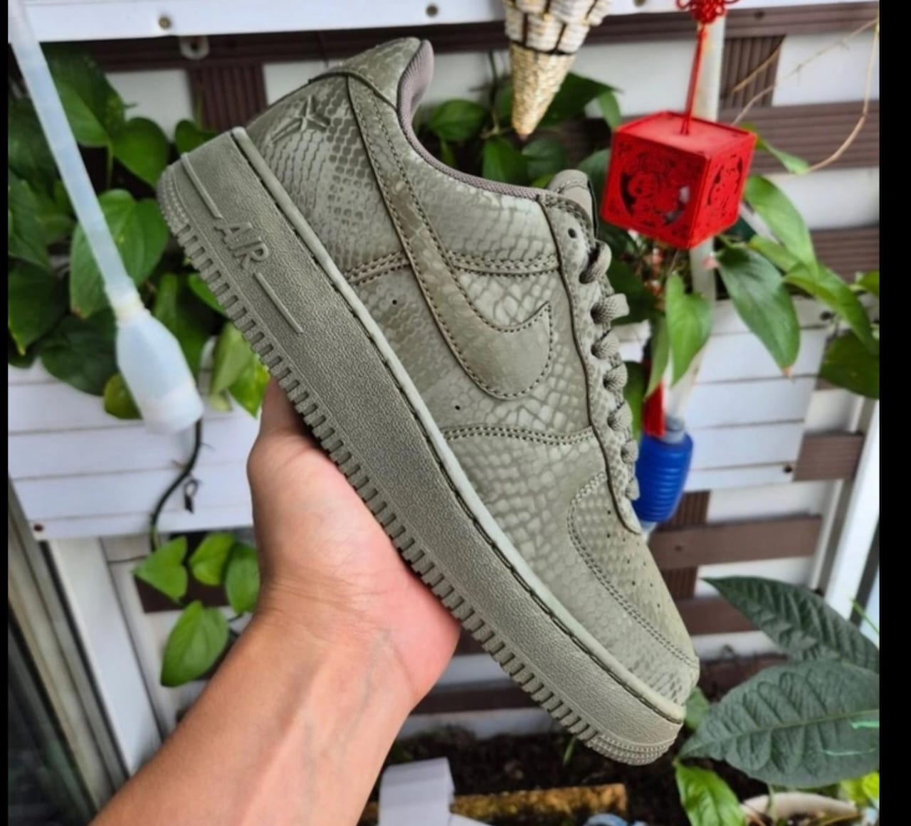 Olive green snakeskin Nike Air Force 1 sneaker held in hand with plants in background.