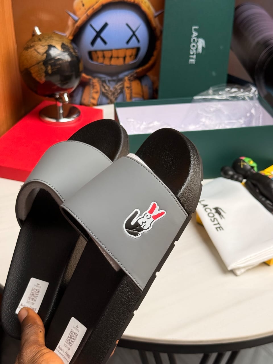 Gray Lacoste slides held in hand showing logo and branding.