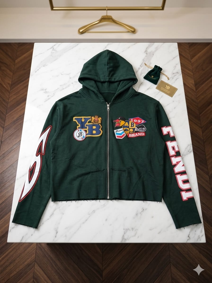 Dark green cropped zip hoodie with YB patches, bird mascot and sleeve prints laid flat on marble.