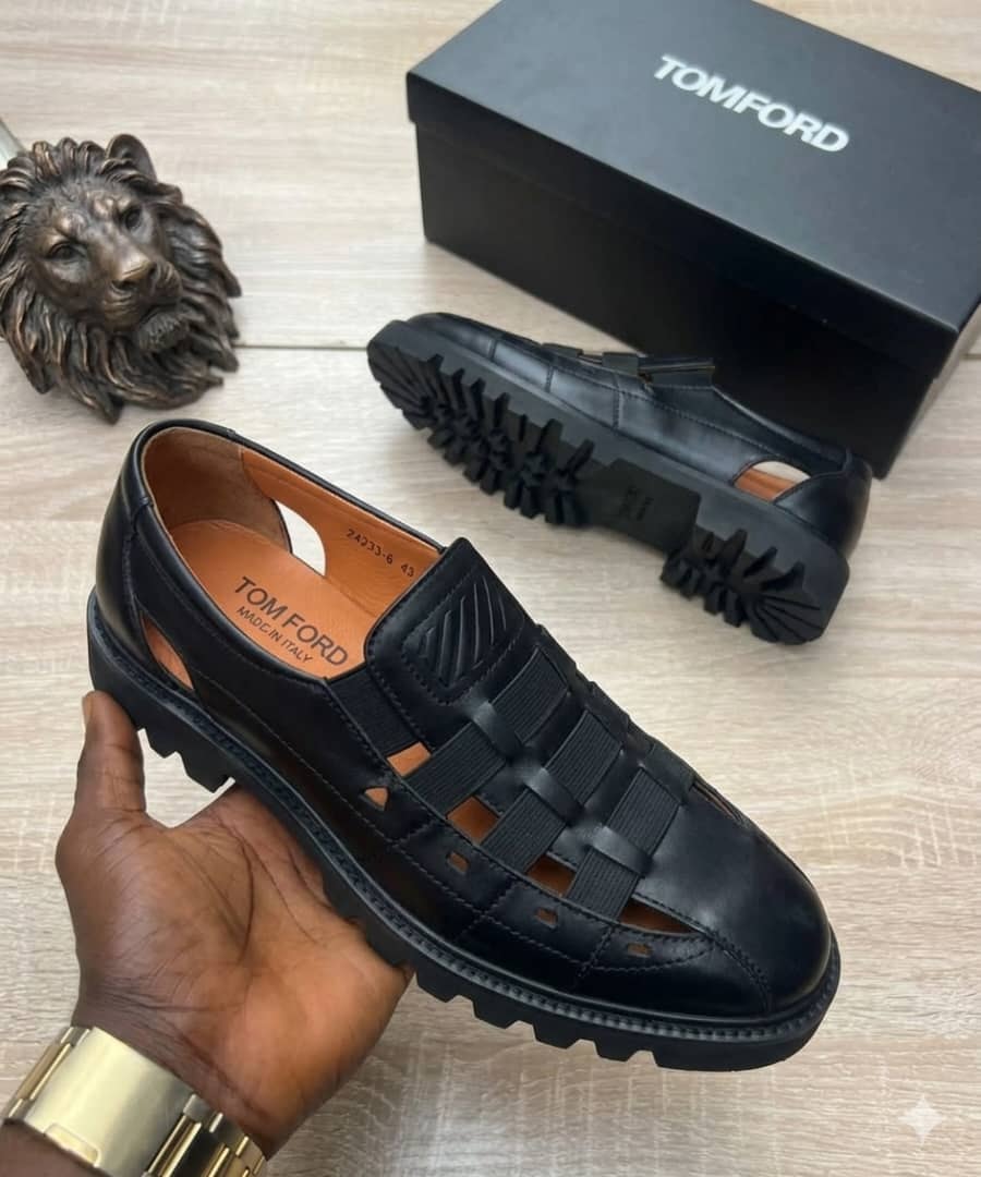 Black Tom Ford loafer with woven straps and cut-outs held in hand beside black branded box.
