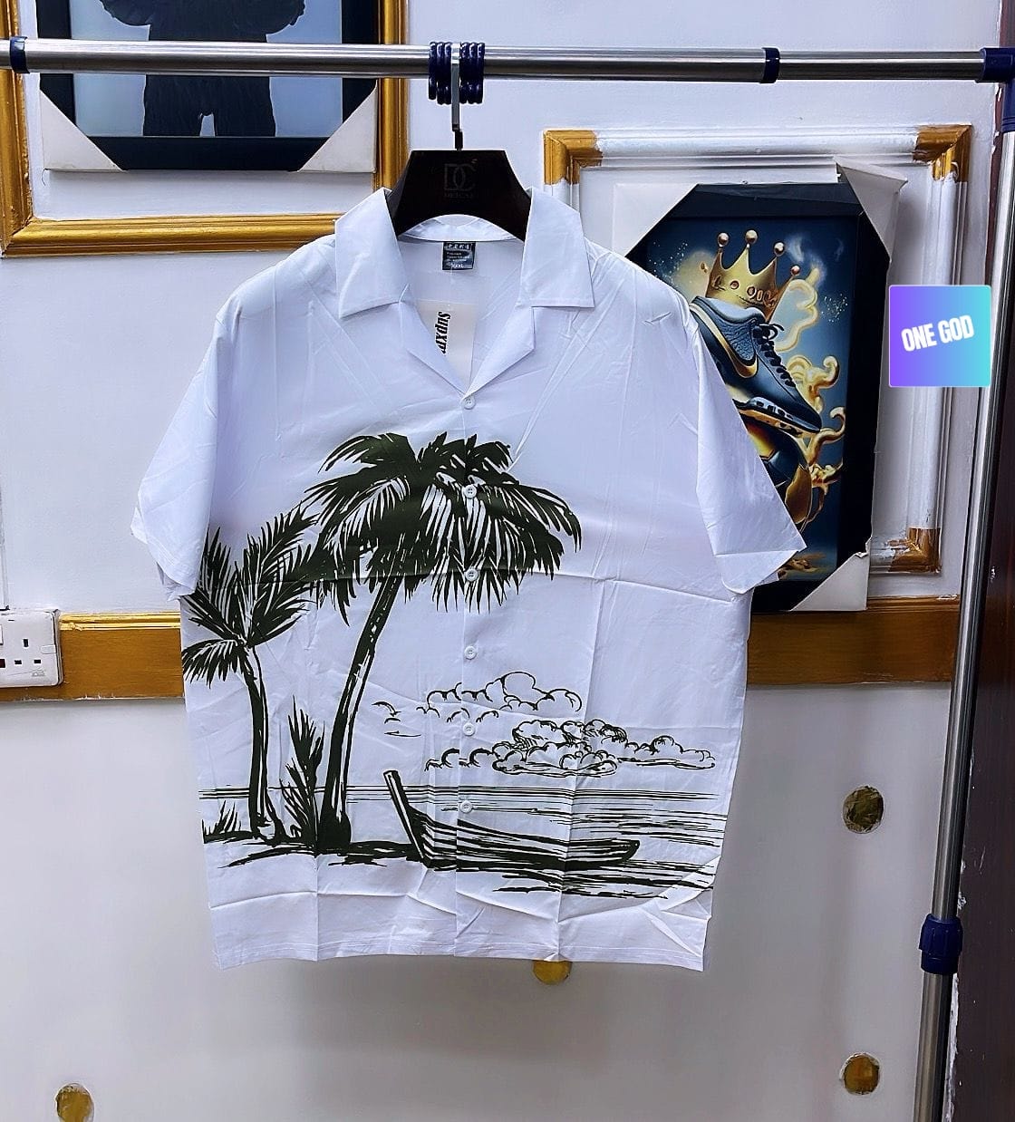 White short sleeve shirt with palm trees and beach scene hanging on rack.