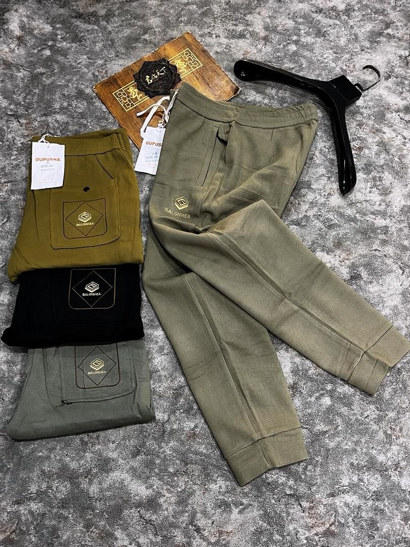 Light olive joggers laid out beside folded pairs in mustard, black and sage with visible branding and tags.