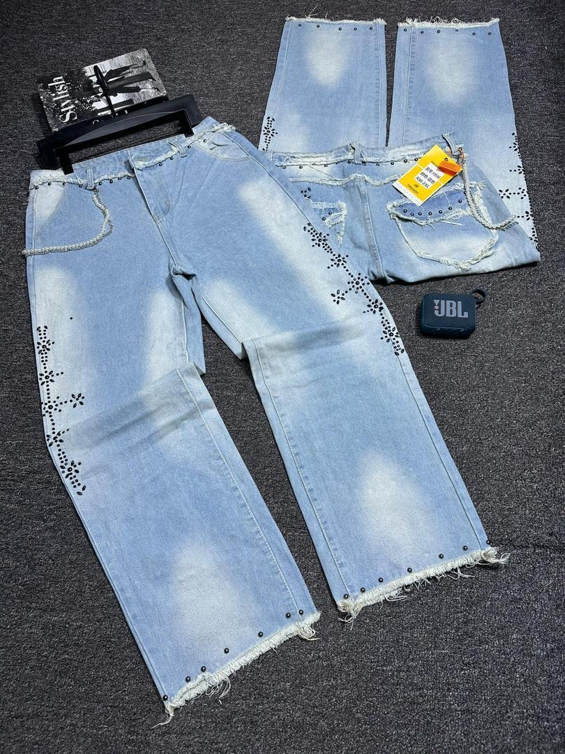 Light blue baggy jeans with stud patterns, rips, and frayed hems shown in flat lay with accessories.