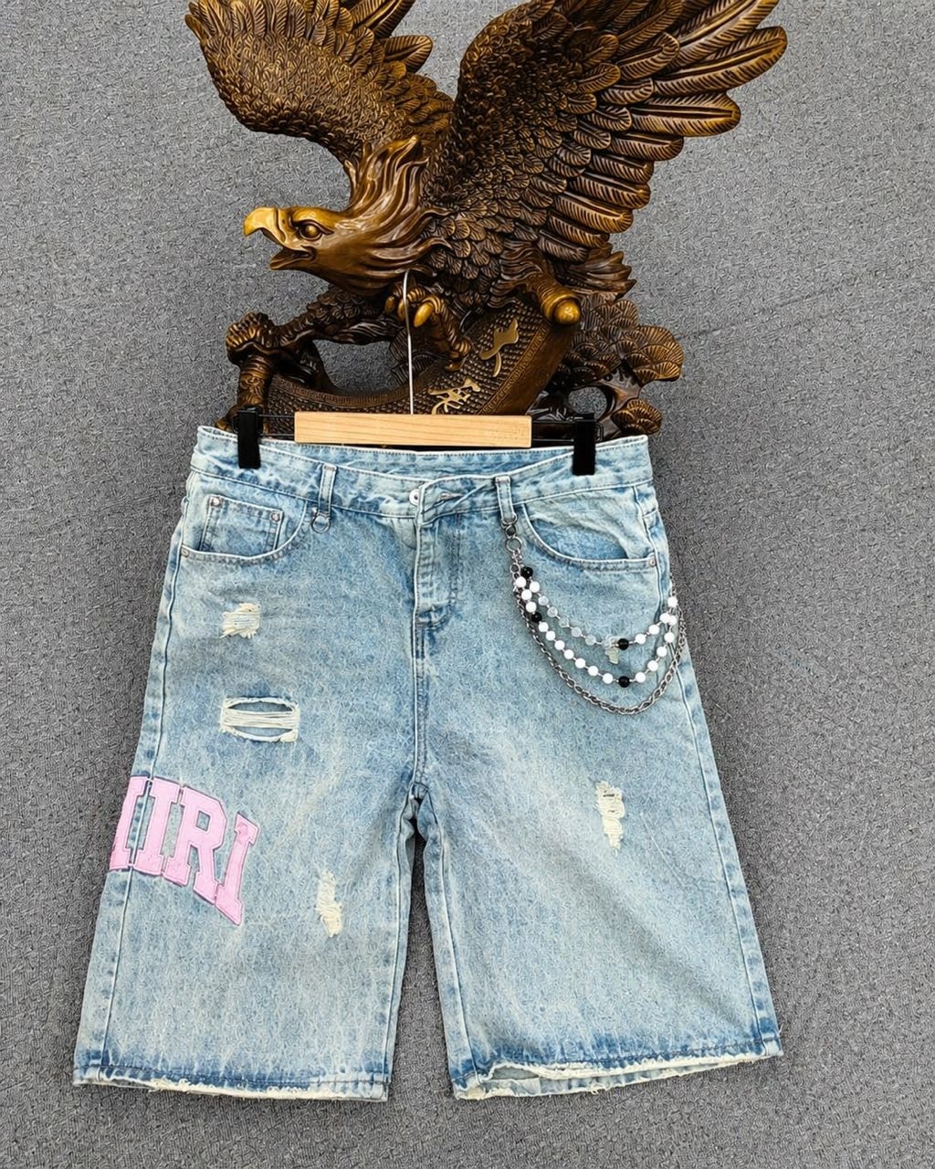 Light blue baggy AMIRI jorts on wooden hanger with pink lettering, distressing, and beaded chain accessory.