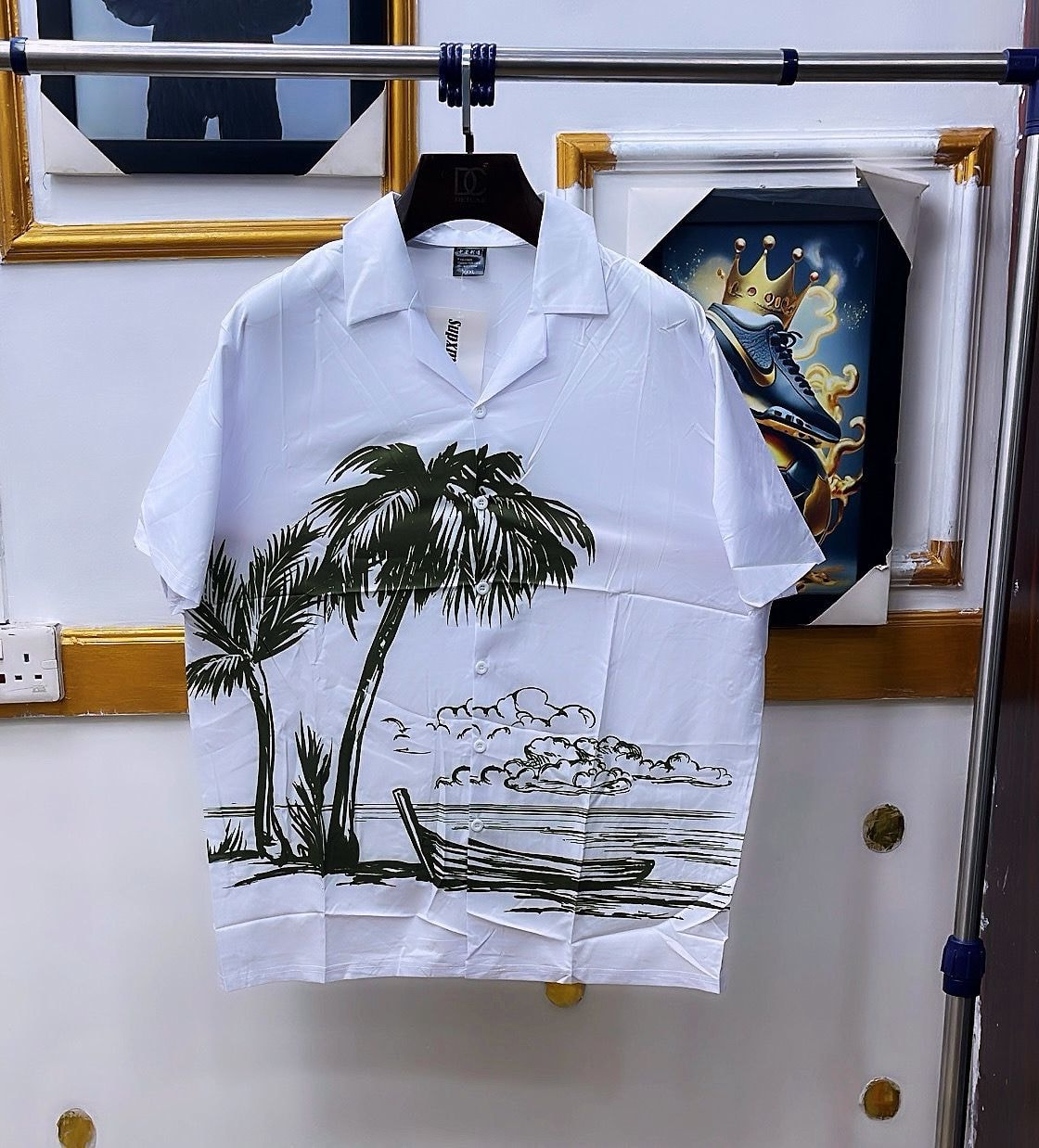 White short sleeve shirt with black palm tree and beach print hanging on rack.