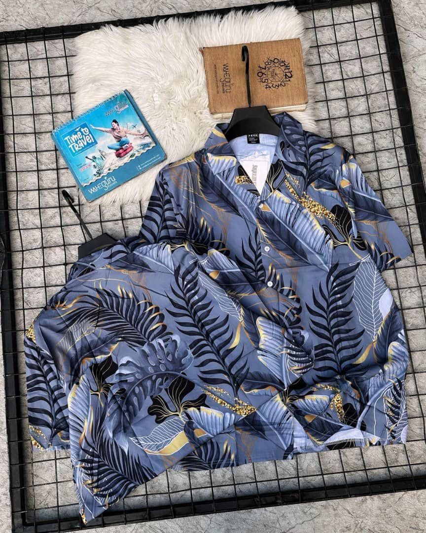 Blue short sleeve shirt with gold and black tropical leaf print laid flat on grid with props.