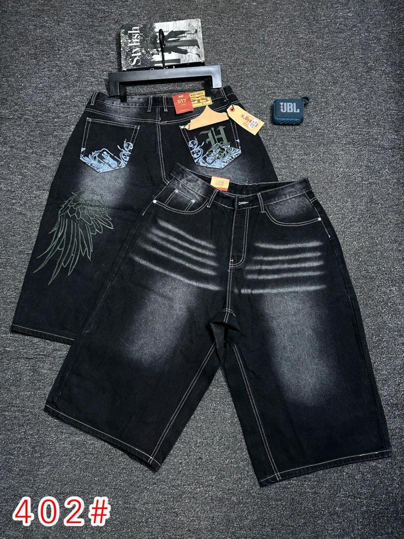 Two pairs of black denim jorts on gray surface, one with prominent wing embroidery on leg and graphic back pockets, the other with horizontal white stripes.