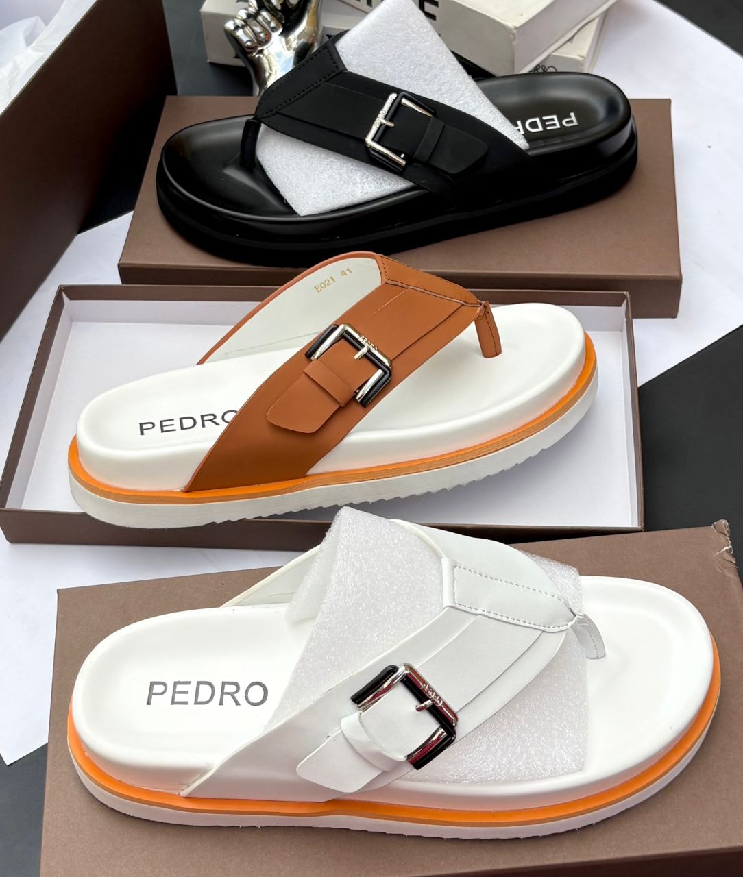Assorted black, brown, and white PEDRO buckle thong sandals displayed in packaging.