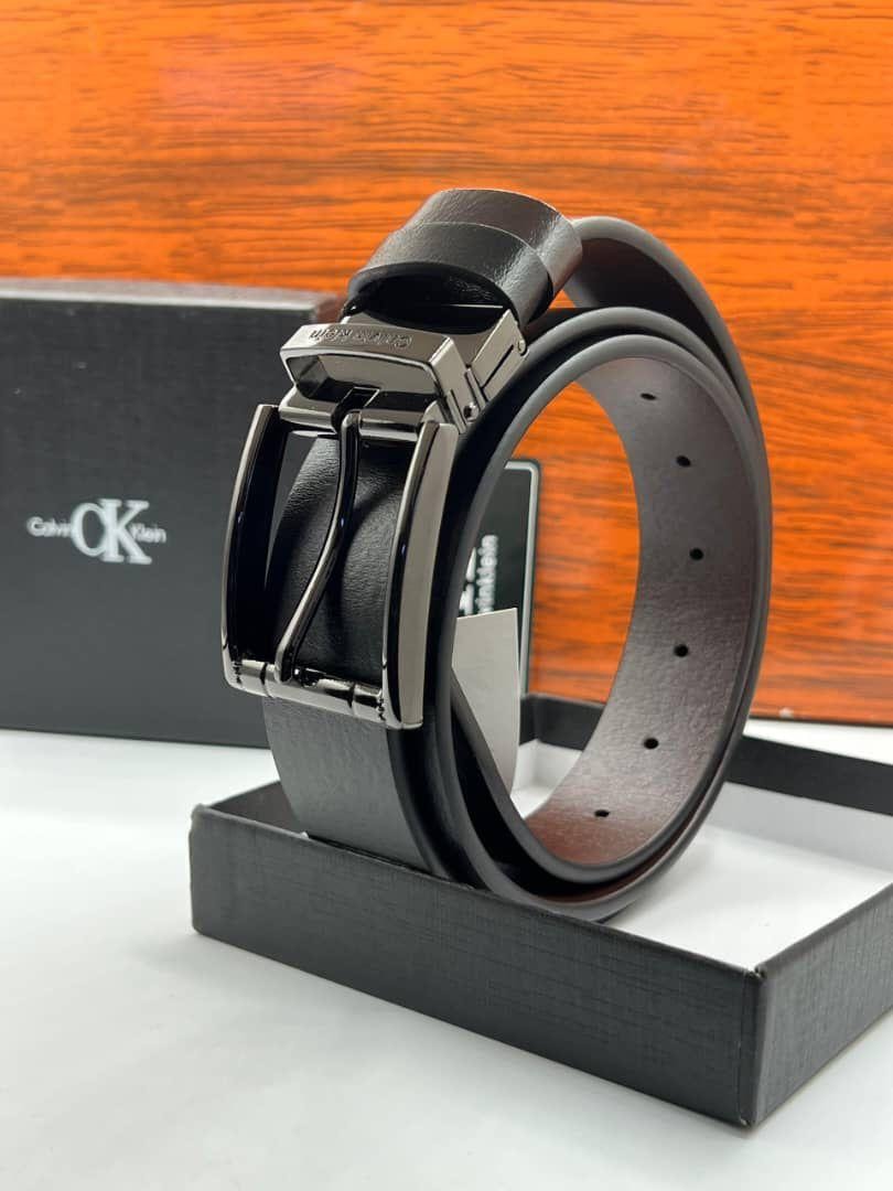 Black Calvin Klein leather belt with silver buckle, tag, and branded black box on wooden surface.