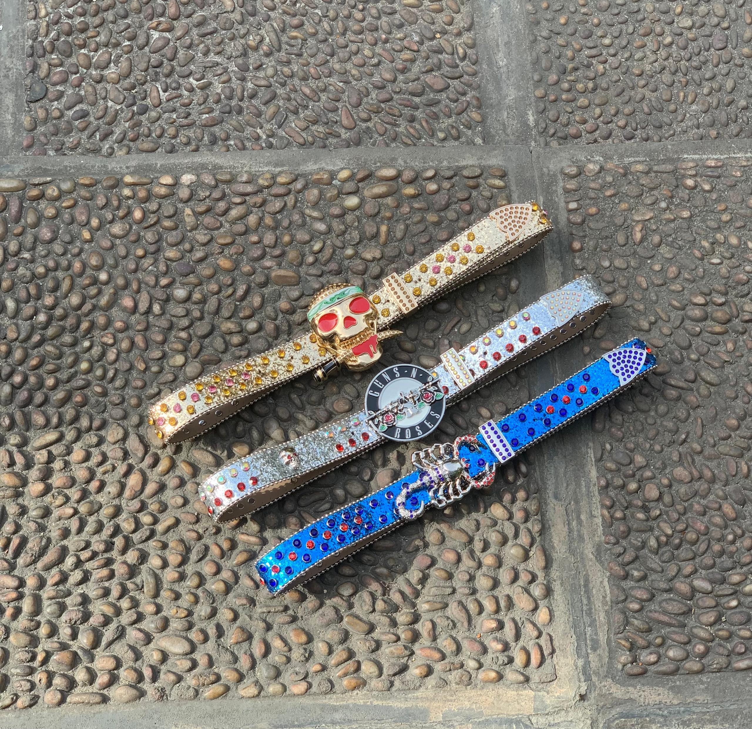 Three rhinestone designer belts with skull and statement buckles in gold, silver and blue on cobblestone.