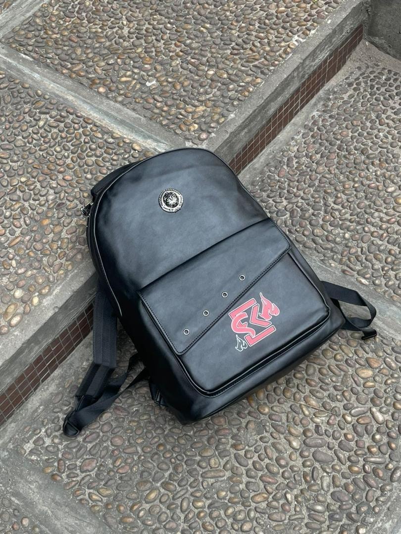 Black backpack with circular silver medallion and red flame logo on cobblestone surface.