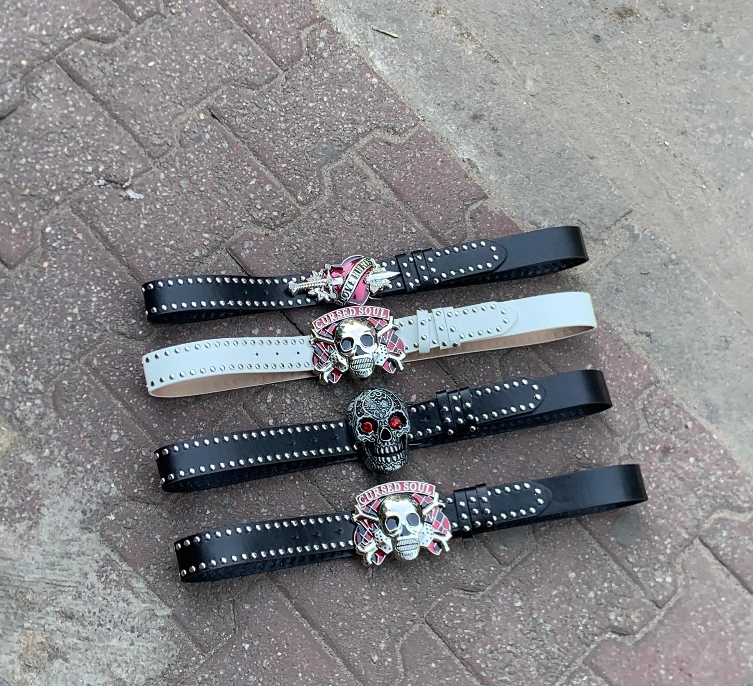Four studded belts with cursed soul skull buckles in black and white on pavement.
