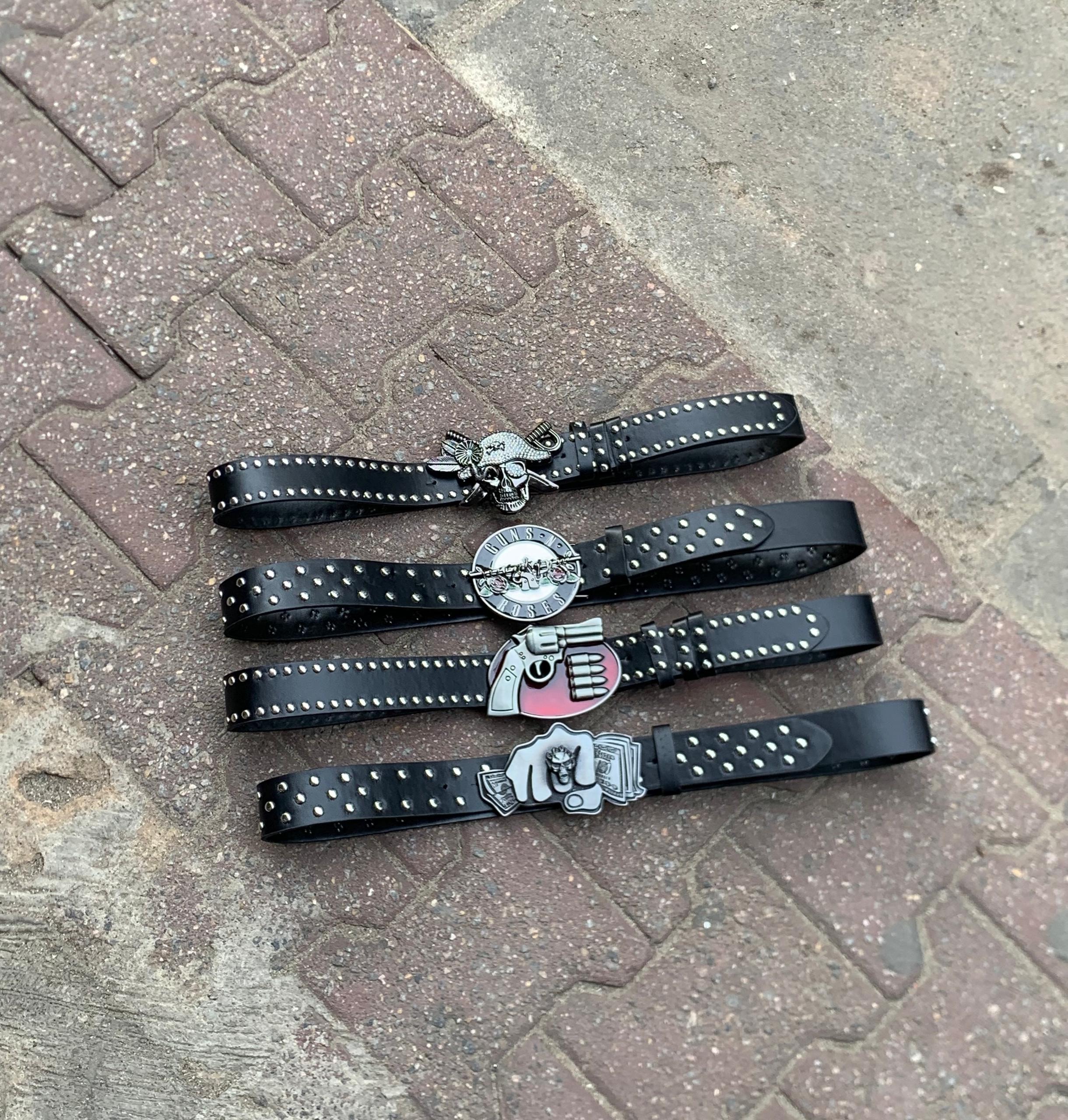 Four black studded leather belts with different statement buckles including skulls, guns, and roses displayed on brick ground.